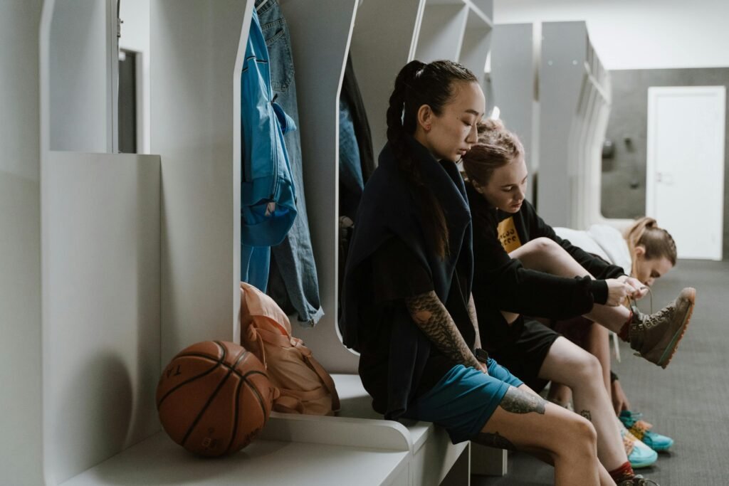 Women basketball players preparing in a locker room, tying shoes, with sports gear around.